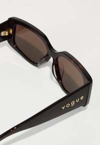 Suknelės iš torto šešiakampio saulės akiniai su tamsiais lęšiais, turintys storą kampuotą rėmelį ir auksinį "vogue" logotipą ant šventyklos. Lygus, blizgus paviršius.