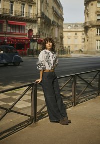 White blouse with black floral embroidery, wide-leg dark denim pants, and brown belt. Standing against a decorative metal railing.