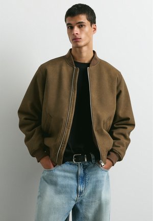 Un homme porte un blouson bomber marron zippé sur une chemise noire et un jean bleu clair, debout les mains dans les poches.
