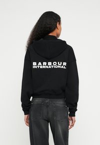 Sweatshirt preta com capuz e a inscrição "BARBOUR INTERNATIONAL" em grandes letras brancas nas costas. Apresenta capuz com cordões e punhos canelados. Combinada com jeans cinza escuro.