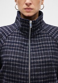Ζακέτα με υψηλό γιακά και φερμουάρ σε ναυτικό μπλε και γκρι καρό σχέδιο. Μαλακό, υφασμάτινο υλικό με ασημένια λεπτομέρεια στο φερμουάρ και ραγαλόν μανίκια.