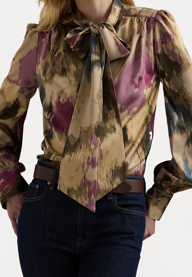 Žena oblečená v blůze s multikolovým abstraktním vzorem a velkým límcem s mašlí, spárovaná s tmavými džíny a hnědým opaskem.