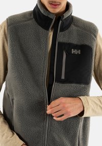 Gilet in pile grigio con tasca frontale in nylon nero. Presenta un colletto alto, chiusura con zip e una superficie morbida e texture.