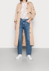 Trench beige con botones, usado sobre una camiseta de tirantes de color claro con efecto tie-dye, combinado con jeans de tiro alto azules y zapatillas blancas.