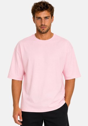 Uomo in piedi contro uno sfondo semplice, indossa una maglietta larga rosa chiaro e pantaloni neri, rivolto in avanti con un'espressione neutra.