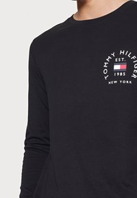 Musta pitkähihainen t-paita, joka on valmistettu puuvillasta ja jossa on pyöreä logo valkoisena sinisillä ja punaisilla yksityiskohdilla rintakehällä.