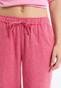 Czerwone i białe spodnie w kratę psyją (houndstooth) z elastycznym pasem i sznurkiem do wiązania, z dwoma kieszeniami po bokach. Wykonane z miękkiej bawełny.