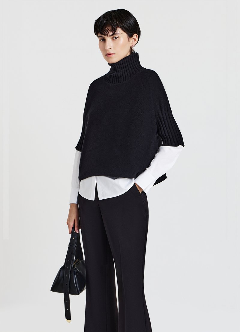 Schwarzer gestrickter Poncho mit hohem Rippkragen, über einem weißen Hemd getragen, kombiniert mit weit geschnittenen schwarzen Hosen und einer kleinen schwarzen Tasche.