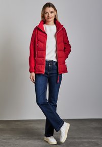 Röd vadderad jacka med huva, med en framdragkedja och dragsko, bärs över en vit tröja och blå jeans, tillsammans med vita sneakers.