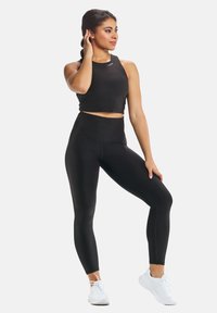 Musta urheilullinen crop-top ja korkeavyötäröiset leggingsit, jotka on valmistettu venyvästä materiaalista. Malli, joka on pukeutunut valkoisiin lenkkarit, seisoo yksi käsi lanteellaan.