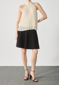 Top halter di un crema pallido con pois neri e plissettature, abbinato a una gonna nera svasata e sandali neri con cinturini su uno sfondo grigio.