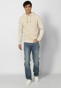 Haut à capuche en tricot beige avec un cordon de serrage, porté avec un jean bleu clair et des baskets blanches. Design simple avec des poignets et un ourlet côtelés.