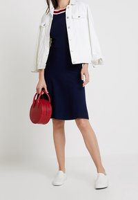 Mujer con vestido azul marino con detalle en el cuello rojo y blanco, chaqueta blanca apoyada sobre los hombros, zapatos blancos de tipo slip-on, sosteniendo un bolso redondo rojo.