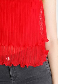 Haut plissé rouge avec une finition texturée, présentant un ourlet ondulé et un design en couches. Associé à un tissu gris foncé. Pas de quincaillerie visible.