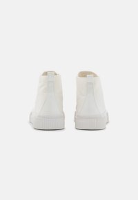 Een paar witte hoge canvas sneakers, gezien vanuit de achterkant op een duidelijke licht achtergrond.