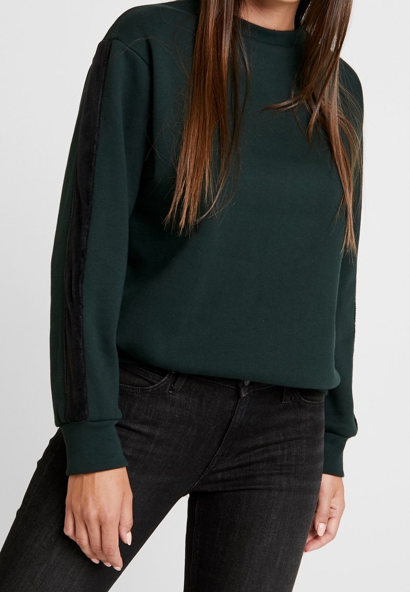 Femme portant un sweat-shirt vert foncé avec une bande noire sur les manches et un jean noir, sur un fond clair uni.