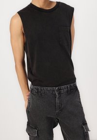 Černý bavlněný top bez rukávů s přední kapsou. Kombinován s tmavě šedými kalhotami typu cargo s několika kapsami a kontrastním šitím.