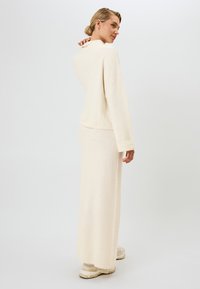 Kermansävyinen neulepusero rentolla istuvuudella ja pitkillä hihoilla, yhdistetty samansävyiseen pitkässä hameeseen. Ominaisuuksina pehmeä tekstuuri ja minimalistinen muotoilu.