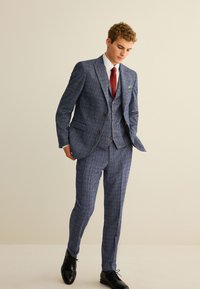 Blauer dreiteiliger Anzug mit Muster, einem Reverskragen, passenden Hosen und einem weißen Hemd. Mit braunen Knöpfen und roter Krawatte. Schwarze Schuhe.