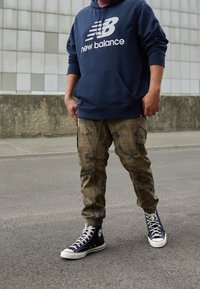 Henkilö, jolla on yllään tummansininen New Balancen huppari, saharanvihreät maastokuosiset cargo-housut ja mustat Converse-korkeavartiset tennarit, seisoo asvaltilla laattaperustaisen seinän vieressä.