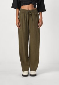 Pantalons larges de couleur vert olive en tissu texturé, avec une ceinture élastique et un cordon de serrage. Associés à des chaussures de couleur crème.