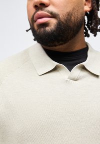 Άνθρωπος με γενειάδα και πλεγμένα μαλλιά, φοράει μπεζ πόλο με υφή πάνω από μαύρο τοπ με στρογγυλή λαιμόκοψη, εμφανιζόμενος από τον λαιμό μέχρι το στήθος.