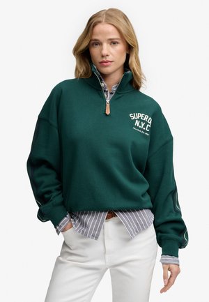 Vrouw draagt een donkergroene quarter-zip sweatshirt met de tekst "SUPERDRY N.Y.C", laag over een gestreept overhemd en witte broek tegen een witte achtergrond.