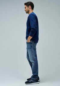 Sudadera navy con puños acanalados, jeans de mezclilla claros y zapatillas negras. El modelo está de lado contra un fondo gris claro.
