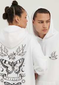 Hvit hettejakker har trykte svarte design, inkludert en sommerfugl, roser og fugler, med et banner som viser teksten "DIN TUR" på baksiden.
