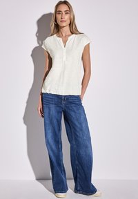 Top corto bianco con maniche corte e una patta di bottoni, abbinato a jeans larghi in denim blu e sneakers bianche su uno sfondo neutro.