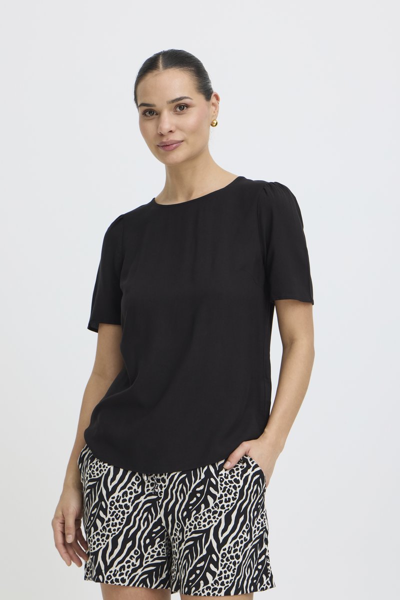 Kobieta z włosami spiętymi do tyłu, ubrana w czarną bluzkę z krótkim rękawem i czarno-białe szorty w zwierzęcy wzór, stojąca z jedną ręką w kieszeni.