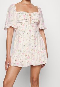 Vestido floral con mangas cortas con volantes, escote cuadrado y parte delantera con cordón. Tejido suave en rosa claro con estampado floral en verde y azul.