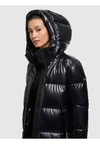 Sjömansblå vadderad jacka med glänsande yta, stor huva, dragkedja fram och tryckknappar. Har vadderade sektioner och en logotypmärke på ärmen.