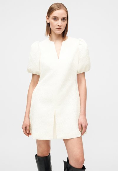 Vestido curto branco texturizado com mangas bufantes, decote em V profundo e uma racha frontal. Usado com botas pretas até ao joelho. Design simples e moderno.