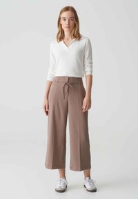 Witte longsleeve top gecombineerd met hoogsmokkel, brede bruine broek met een trekkoord. Model staat in witte sneakers op een effen achtergrond.