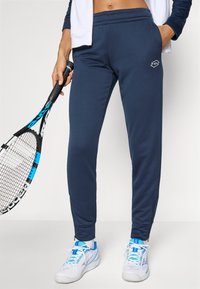 Jūrinės sportinės kelnės su raišteliu, liekna figūra, šoninės užtrauktukų kišenės, subtilus logotipo detalė, derinamos su baltomis sportinėmis bateliais.