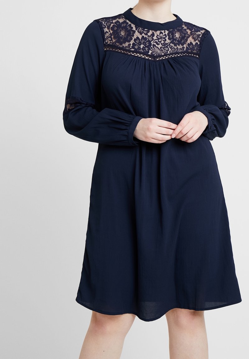 Femme portant une robe bleu marine à manches longues, avec des détails en dentelle sur le haut de la poitrine et les manches, debout devant un fond uni.