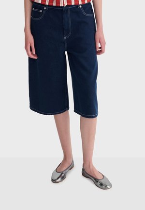 Person trägt dunkelblaue, kniehohe Jeansshorts, ein rot-weiß gestreiftes Hemd und metallisch silberne Ballerinas und steht auf einem grauen Boden.
