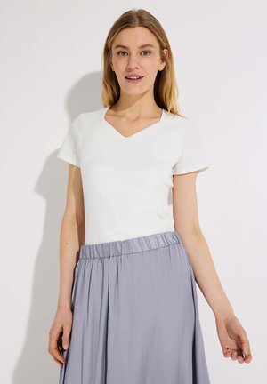 Junge Frau mit langen blonden Haaren, die ein weißes T-Shirt mit V-Ausschnitt und kurzen Ärmeln sowie einen hellgrauen Faltenrock trägt, steht vor einem weißen Hintergrund.