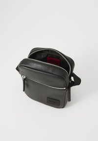 Svart textured skulderveske med glidelås, med en frontlomme og rødt logofôr. Laget av slitesterkt materiale med justerbar stropp.