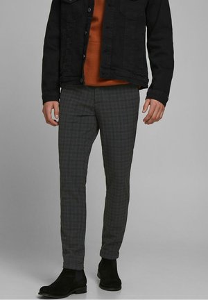 Mann trägt graue karierte Slim-Fit-Hose, schwarze Wildleder-Chelsea-Stiefel, rostfarbenen Pullover und schwarze Denimjacke vor grauem Hintergrund.