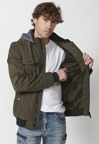 Giacca verde oliva con cappuccio, chiusura con zip e un morbido rivestimento in pelliccia sintetica. Presenta tasche e polsini a coste per un maggior calore.