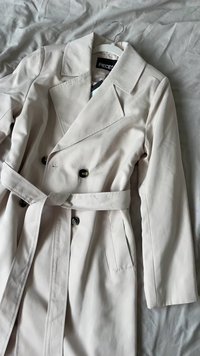 Bež trench kaput napravljen od glatkog materijala. Dvostruko kopčanje s crnim dugmadi, širokim oštrim reverima i odgovarajućim remenom oko struka.