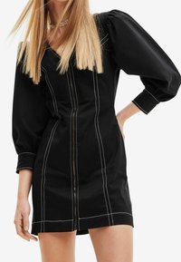 Robe noire avec manches bouffantes, couture blanche et fermeture éclair à l'avant. Présente un buste ajusté et une coupe droite, fabriquée en tissu de mélange de coton.