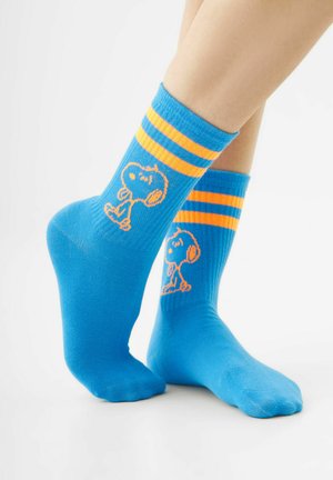 Blaue Sportsocken mit orangefarbenen Streifen und einem Cartoon-Hundmotiv, getragen an den Füßen vor weißem Hintergrund.