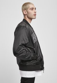 Chaqueta bomber negra con un acabado brillante, que presenta un cierre frontal, puños acanalados y un audaz logo blanco en el pecho. Textura suave, ajuste casual.