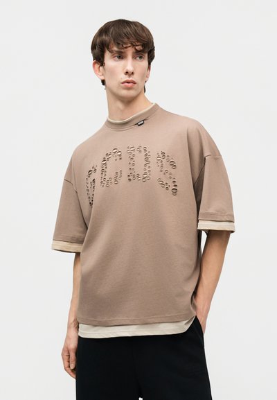 Νεαρός άνδρας που φοράει ένα φαρδύ καφέ μπεζ T-shirt με διάτρητα γράμματα στο στήθος, φορεμένο πάνω από ένα μπεζ εσώρουχο, στέκεται μπροστά σε απλό φόντο.