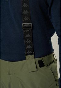 Zielone wodoodporne spodnie z czarnymi regulowanymi szelkami; posiadają teksturowaną dzianinową bluzkę w kolorze granatowym; zawierają czarne akcenty metalowe.