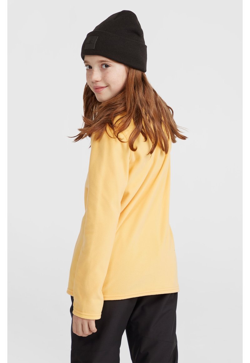 Gelbes, langärmeliges Fleece-Shirt mit gerippten Bündchen, kombiniert mit einer schwarzen Strickmütze. Naturhaar fällt über die Schultern, vor einem neutralen Hintergrund.