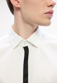 Witte overhemd met een klassieke kraag, voorzien van een zwarte gestructureerde strook aan de voorkant. Knopdetail bij de hals, katoenen stof, gladde textuur.